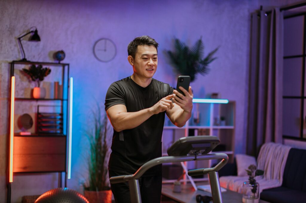 Attractive asian man with smartphone exercising on treadmill at home  gym at evening time.