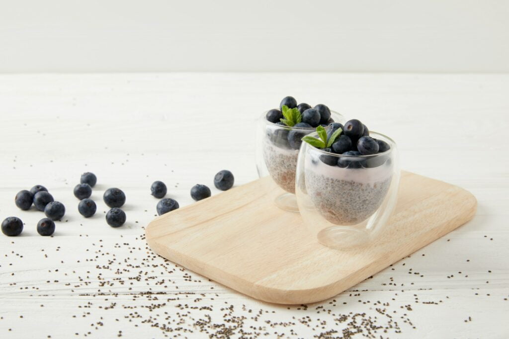 close up view of chia puddings with fresh blueberries and mint on wooden cutting board on white