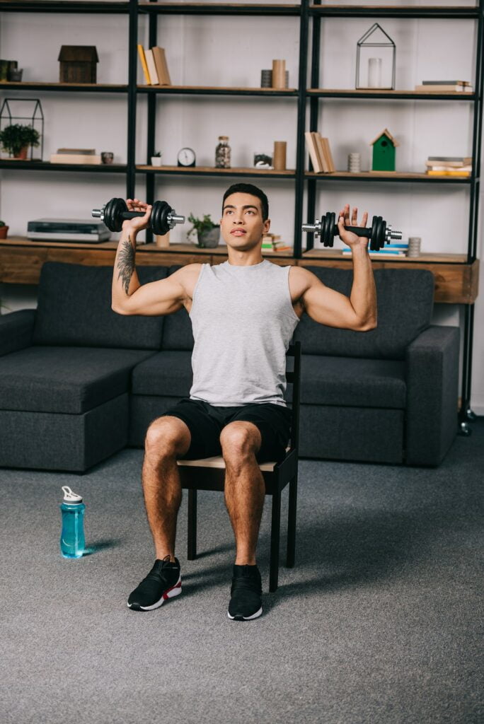 tattooed mixed race man exercising with dumbbells on chair in home gym