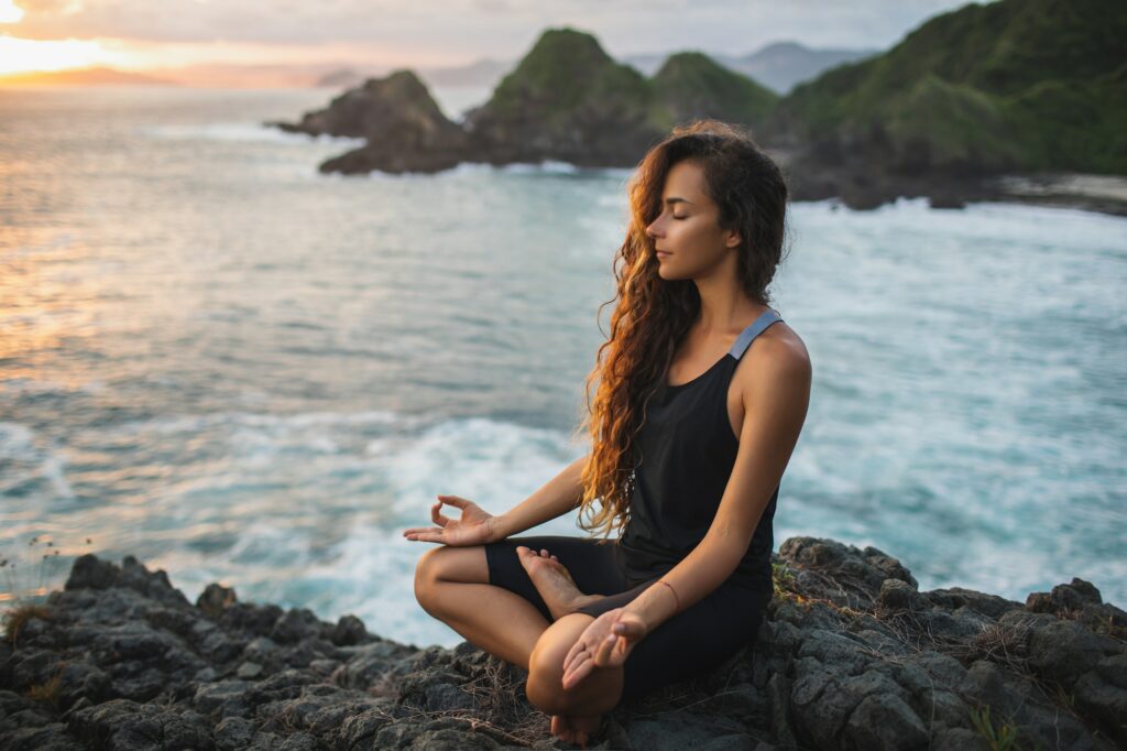 Young woman practicing yoga in lotus pose at sunset with beautiful ocean and mountain view